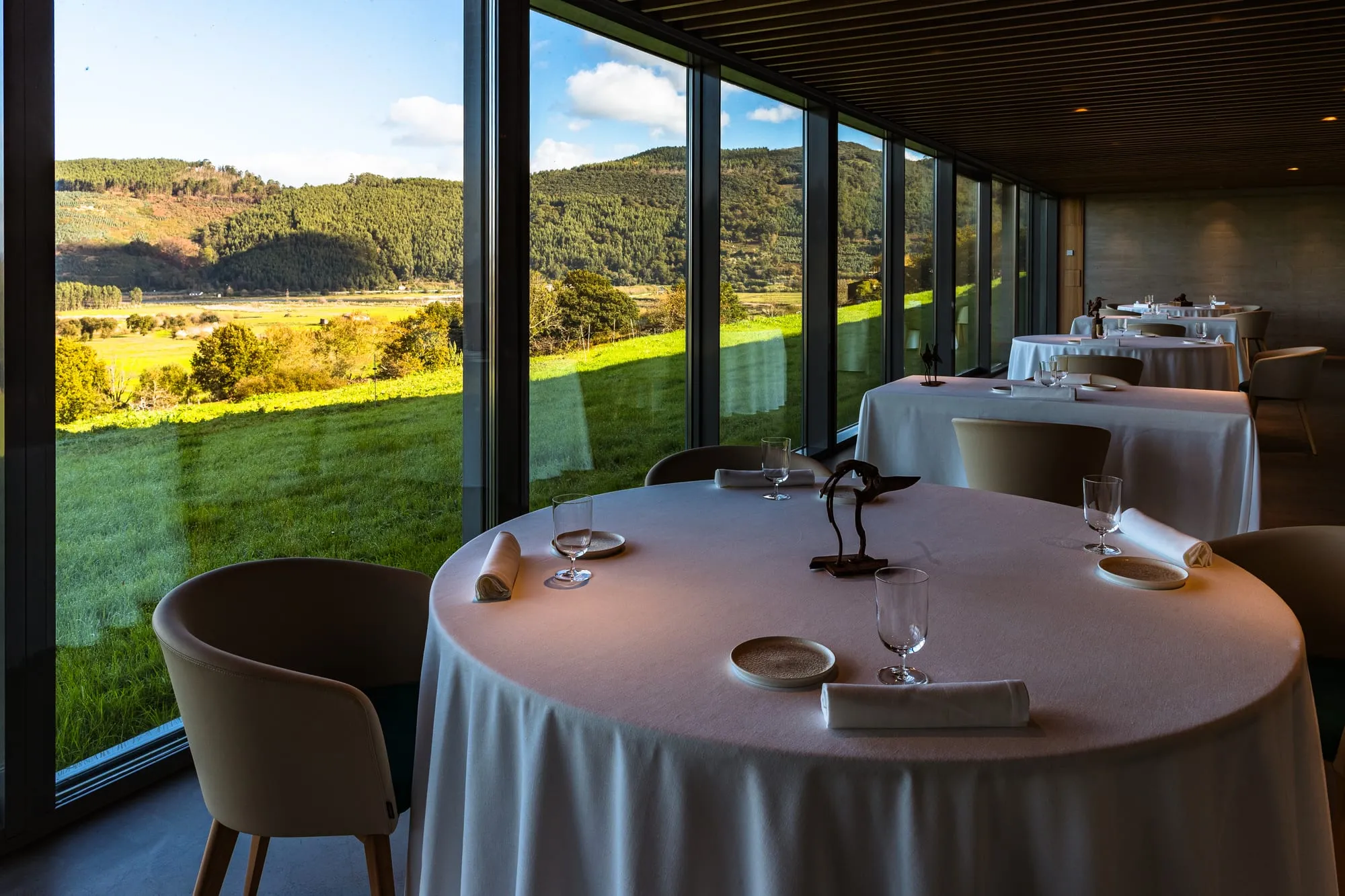 Mesa del comedor con mantel blanco junto al ventanal, con los prados y las montañas al atardecer