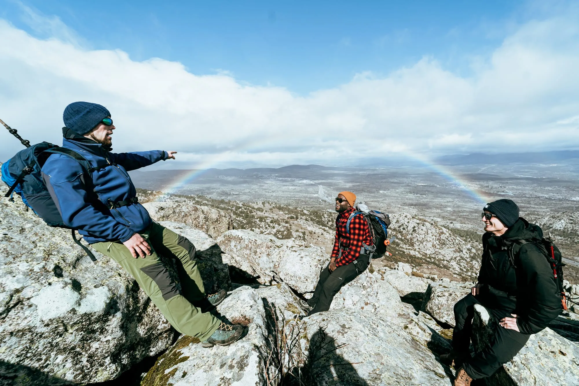Tres senderistas sentados en las rocas de una cumbre con un arcoíris sobre el valle