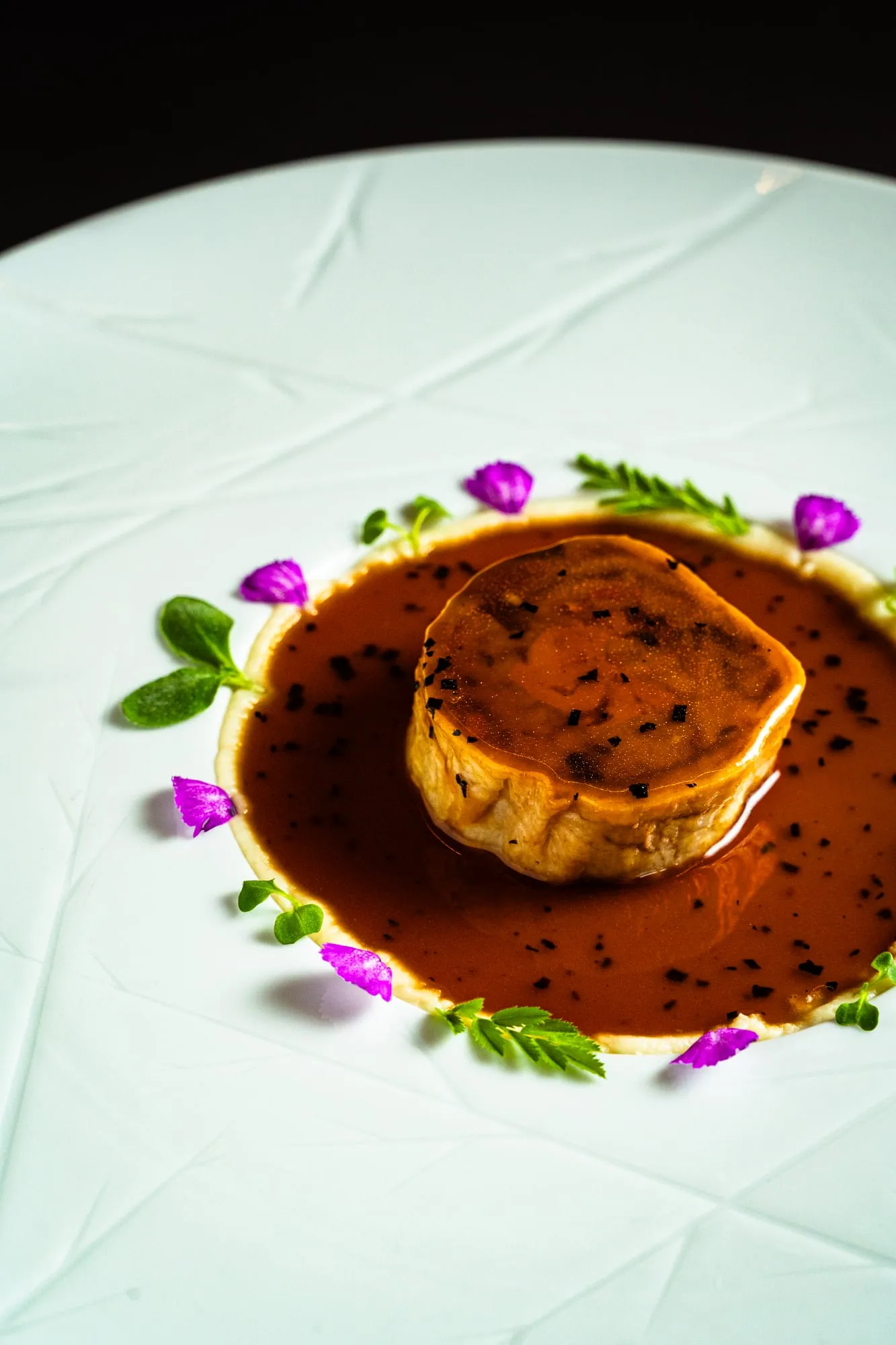 Pieza glaseada sobre salsa oscura con flores y hojas en un plato blanco texturizado