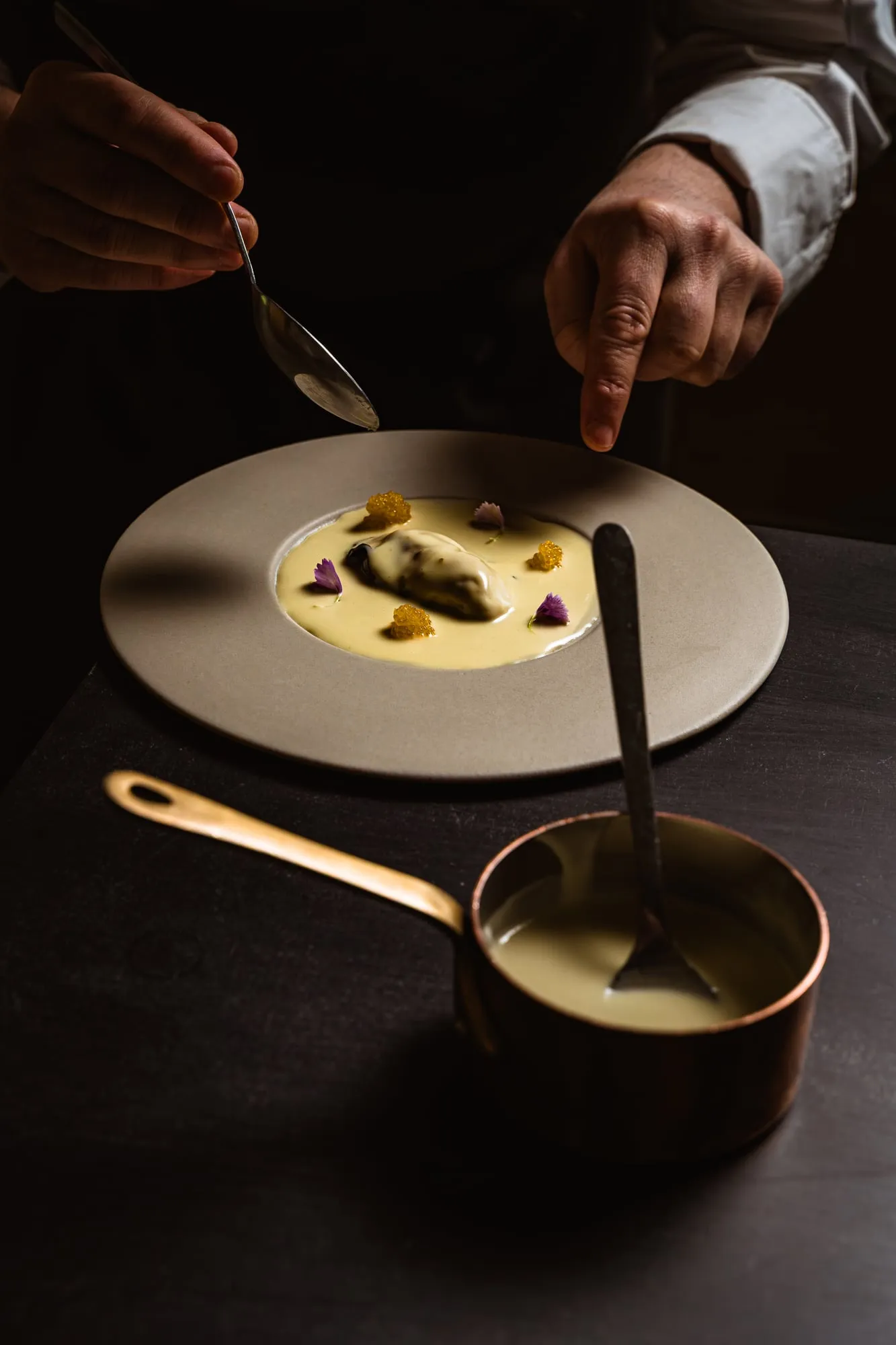 Manos del equipo de cocina terminando un plato con salsa junto a un cazo de cobre