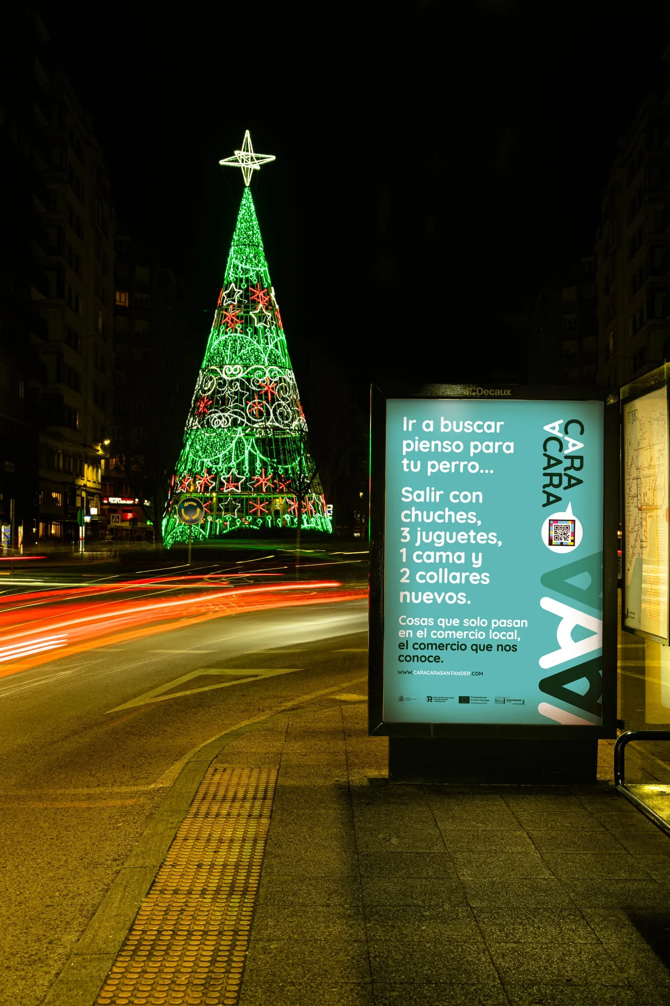 Mupi de Cara a Cara iluminado de noche junto al árbol de Navidad de luces verdes