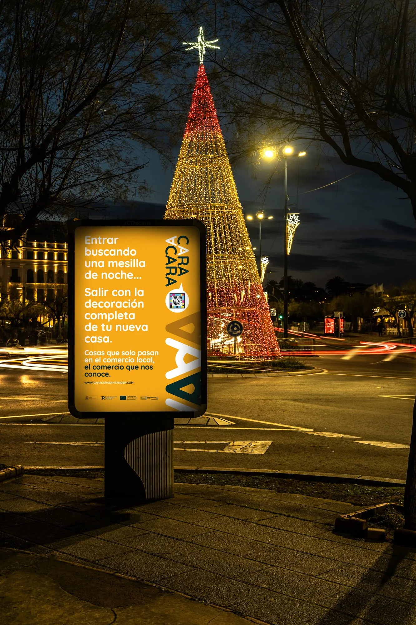 Mupi naranja de la campaña de noche frente a un gran árbol de Navidad iluminado